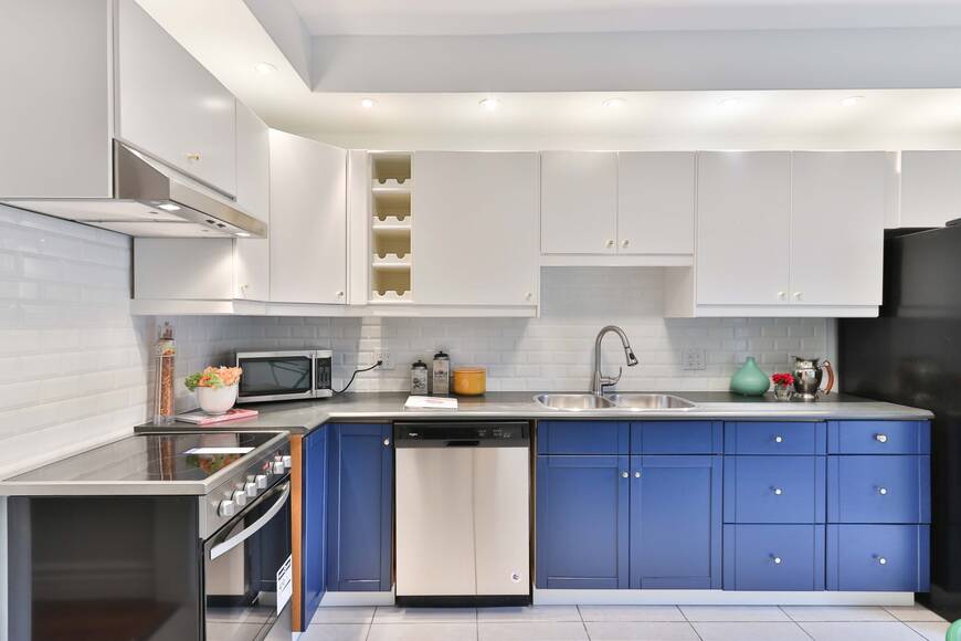 Stylish modular kitchen by Alea Modular featuring two-tone cabinetry with blue lower cabinets and white upper cabinets, a sleek subway tile backsplash, and built-in stainless steel appliances. A modern, space-efficient kitchen design with ample storage and elegant finishes.