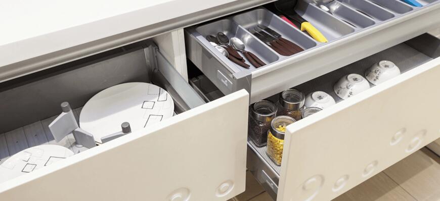 Modern kitchen drawers opened, revealing organized cutlery, cooking utensils, and jars of food alongside round plates.